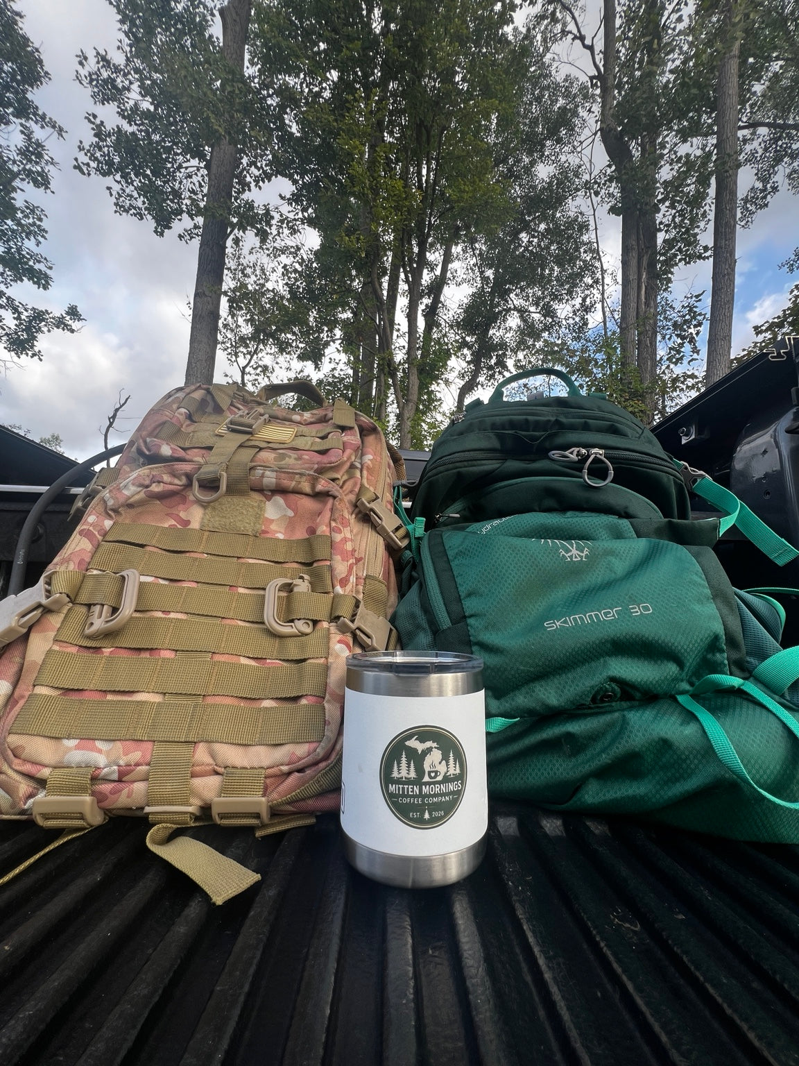 Two outdoor backpacks beside a white Mitten Mornings Coffee Company travel mug in the bed of a truck.